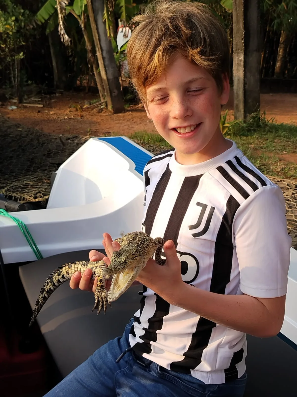 A smiling young guest carefully holding a small crocodile beside a Bentota river safari boat