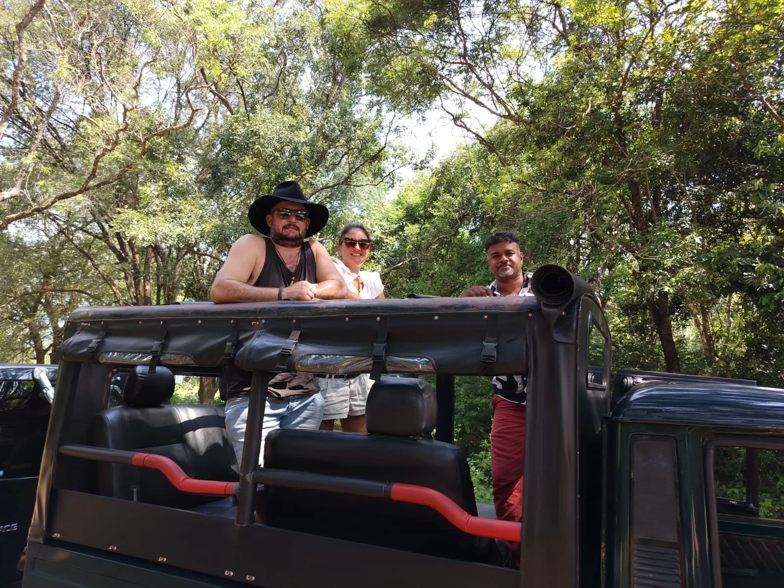 Two guests and their Sammy Tours guide standing together in an open safari jeep beneath forest trees